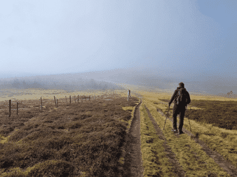 Randonneur avec sac à dos et bâtons sur chemin de terre le long d'une clôture sur le Chemin de Stevenson. Plateau brumeux avec herbe dorée.