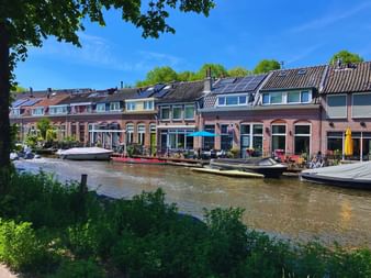 Rangée de maisons colorées le long d'un canal hollandais avec façades en brique rouge. Plusieurs bateaux sont amarrés devant, des arbres verts encadrent la scène.