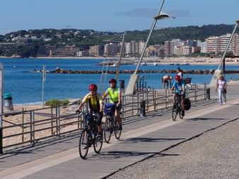 Groupe de cyclistes sur une promenade côtière à Calonge, Costa Brava. Mer Méditerranée bleue, plage et bâtiments sur la colline visibles.