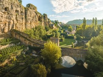 Pont en pierre et remparts anciens à Luxembourg avec falaises spectaculaires, végétation verdoyante et viaduc en arrière-plan.