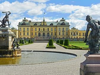 Château de Drottningholm à Stockholm avec façade baroque jaune, jardins à la française, fontaines ornées et statues classiques sous ciel nuageux.