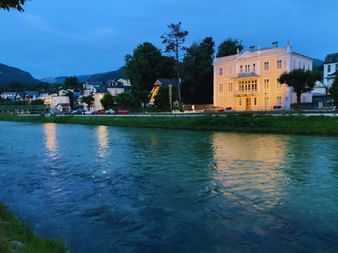 Vue du soir de Bad Ischl avec villa blanche illuminée au bord de la rivière, montagnes en arrière-plan, ciel bleu crépusculaire se reflétant dans l'eau.