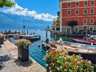 Port pittoresque de Limone avec bateaux colorés amarrés dans l'eau turquoise, jardinières fleuries sur le quai en pierre et bâtiment rouge.