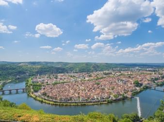 Vue aérienne de Cahors dans la vallée du Lot, montrant le centre historique entouré d'un méandre du Lot avec ponts et collines.