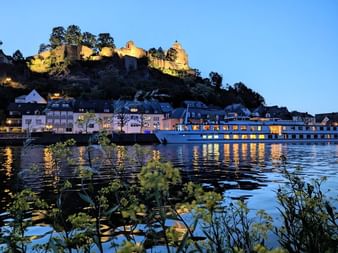 Bateau de croisière MS Olympia amarré au bord de l'eau à Sarrebourg au crépuscule, avec château médiéval illuminé sur la colline et bâtiments traditionnels.