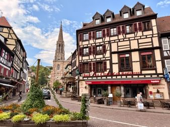 Bâtiments à colombages traditionnels avec volets bruns bordent une place pavée en Alsace. Un clocher s'élève en arrière-plan.