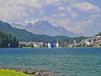 Lac de Saint-Moritz avec eau turquoise, rive verte, voiliers et la ville alpine de Saint-Moritz adossée à des montagnes boisées.