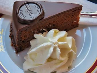 Tranche triangulaire de Sachertorte au chocolat avec glaçage chocolat noir et sceau Hotel Sacher, servie avec crème fouettée décorative.