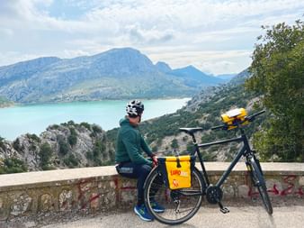 Cycliste en veste verte et casque assis sur mur de pierre surplombant réservoir turquoise entouré de montagnes à Majorque.