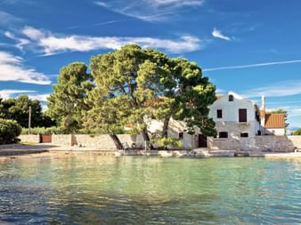 Bâtiment de couvent en pierre blanche avec toit de tuiles rouges sur l'île Ugljan, Croatie, situé près d'eaux turquoise avec de grands pins.