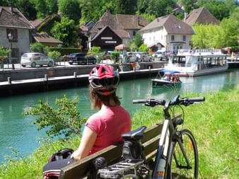 Cycliste en casque et maillot roses assise sur banc en bois au bord du canal turquoise d'Annecy, vélo garé à côté, bateaux sur l'eau.