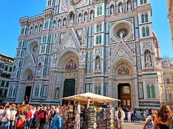 Façade en marbre ornée de la cathédrale de Florence avec motifs verts, blancs et roses. Touristes et stands de souvenirs sur la place.