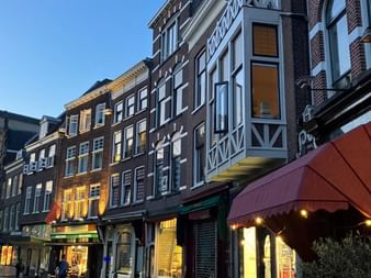 Bâtiments hollandais traditionnels avec façades en brique et colombages blancs bordent une rue pavée au crépuscule. Auvents de magasins et éclairage chaleureux créent une atmosphère accueillante.