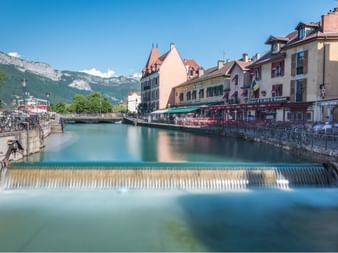 Canal avec cascade dans la vieille ville d'Annecy, bâtiments historiques colorés bordent le cours d'eau avec montagnes en arrière-plan.