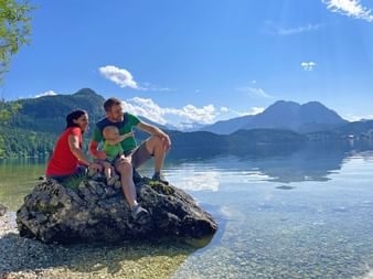 Famille de trois personnes assise sur un gros rocher au bord du lac Altaussee dans le Salzkammergut, Autriche. Montagnes et ciel bleu se reflètent dans l'eau calme.