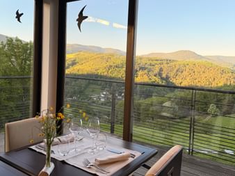 Table de petit-déjeuner dressée avec verres et fleurs devant grandes fenêtres donnant sur montagnes boisées des Hautes-Vosges.