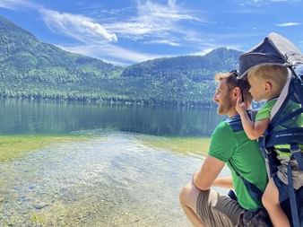 Père en chemise verte portant un enfant dans un porte-bébé au lac Altaussee dans le Salzkammergut, Autriche. Eau cristalline et montagnes boisées.
