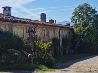 Ancienne ferme jaune avec toit en terre cuite et cheminées à Camigliano, Toscane. Jardin avec fleurs et arbres borde un chemin.
