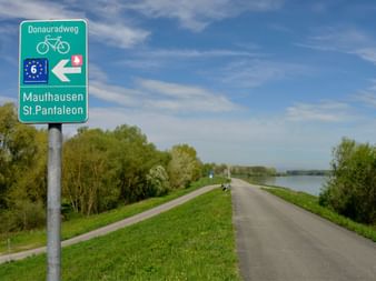 Panneau vert de la piste cyclable du Danube vers Mauthausen et St. Pantaleon, avec route et piste cyclable le long du Danube.