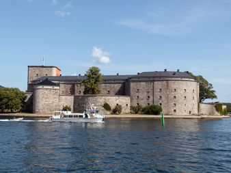 Forteresse Vaxholm avec murs en pierre et tours rondes au bord de l'eau près de Stockholm. Un bateau blanc passe sur l'eau bleue.