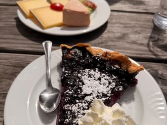 Part de tarte aux myrtilles avec sucre glace et crème fouettée sur une assiette blanche. Un plateau de fromages et charcuterie est visible en arrière-plan.
