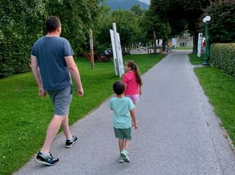 Homme marchant avec deux enfants sur un sentier pavé à travers un parc verdoyant à Bad Ischl, avec des montagnes visibles en arrière-plan sous un ciel nuageux.