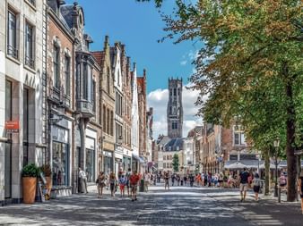Rue pavée à Bruges bordée de bâtiments historiques. Le beffroi emblématique se dresse en arrière-plan. Des piétons se promènent.