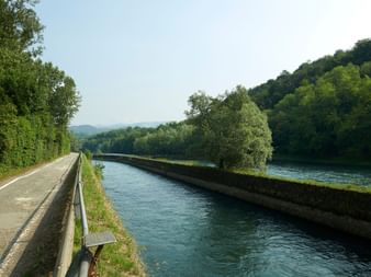 Piste cyclable pavée le long de la rivière Adda bleue avec des collines boisées vertes. Un arbre solitaire se dresse entre le chemin et l'eau.