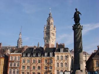 Bâtiments flamands colorés à Lille avec beffroi orné et colonne surmontée d'une statue au premier plan sous un ciel bleu.