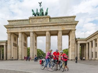 Trois cyclistes avec casques et vêtements colorés debout avec leurs vélos devant la Porte de Brandebourg à Berlin sur une place pavée.