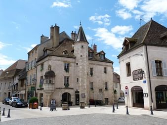 Place de Beaune avec bâtiments historiques en pierre, tourelle, voitures garées et Hôtel Athanor. Ciel bleu avec nuages blancs.