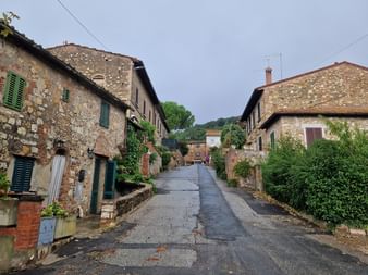 Rue asphaltée mouillée à Iano, Toscane, bordée de maisons en pierre traditionnelles avec volets verts sous un ciel nuageux.