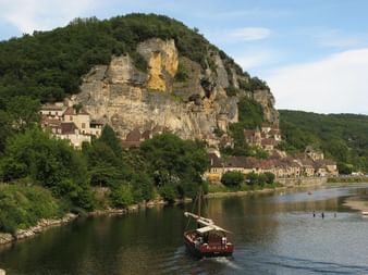 Village de La Roque-Gageac niché contre une falaise calcaire au bord de la Dordogne. Un bateau traditionnel navigue sur l'eau.
