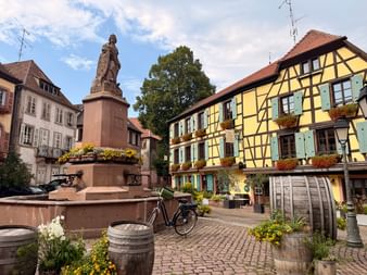 Place de la Route des Vins d'Alsace avec statue en pierre, fontaine, maison à colombages jaune avec volets bleus et tonneaux de vin fleuris.