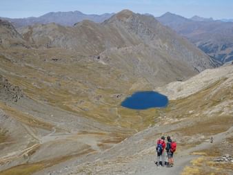 Trois randonneurs avec sacs à dos debout sur une pente de montagne admirant un lac alpin bleu vif entouré de pics arides dans le Queyras.