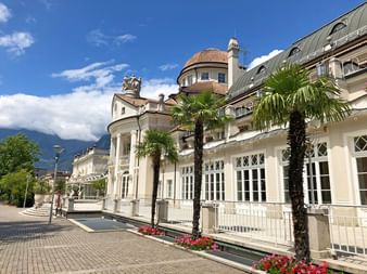 Kurhaus de Merano avec palmiers et montagnes Bâtiment historique Kurhaus à Merano avec façade blanche, toit de tuiles rouges et palmiers. Montagnes visibles en arrière-plan.