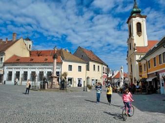 Place pavée à Szentendre avec clocher baroque, bâtiments historiques colorés et personnes dont un enfant à vélo sous ciel bleu.