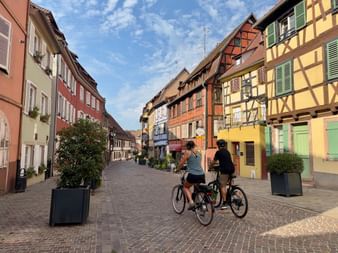 Deux cyclistes sur rue pavée bordée de maisons à colombages colorées en Alsace. Architecture traditionnelle avec façades roses, jaunes et oranges.