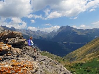 Randonneur en veste violette assis sur affleurement rocheux surplombant vallée montagnarde spectaculaire du Queyras sous ciel nuageux.