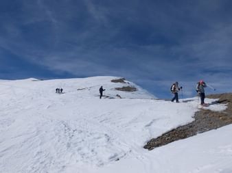 Groupe de raquetteurs avec sacs à dos et bâtons traversant les pentes enneigées du Queyras sous un ciel bleu nuageux.