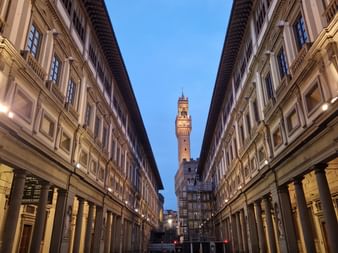 Tour illuminée à Florence au crépuscule, encadrée par des bâtiments Renaissance symétriques avec des colonnades voûtées des deux côtés d'une rue étroite.