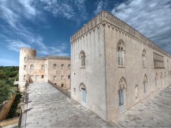 Palais Donnafugata en Sicile avec fenêtres gothiques en arc, façade en pierre et tour crénelée sous un ciel nuageux dramatique.