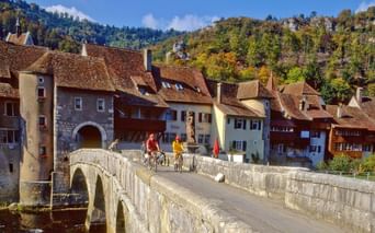 Cyclistes traversant le pont de pierre médiéval sur le Doubs à Saint-Ursanne, avec bâtiments historiques et collines boisées.