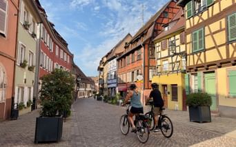 Deux cyclistes sur rue pavée bordée de maisons à colombages colorées en Alsace, France. Architecture traditionnelle aux façades pastel sous ciel bleu.