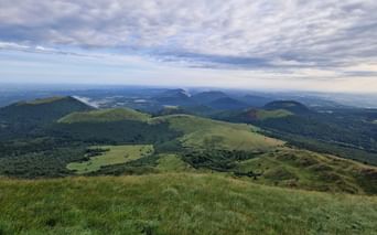 Vue étendue depuis le Puy de Dôme montrant des collines volcaniques vertes et des montagnes s'étendant à l'horizon sous un ciel nuageux.