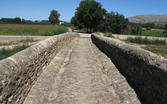 Ancien pont en pierre aux murs patinés traversant le paysage rural de la Costa Brava avec champs, arbres et collines sous ciel bleu.