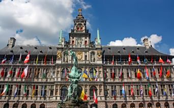 Hôtel de ville historique d'Anvers avec façade Renaissance ornée, tour d'horloge centrale et statue de bronze au premier plan. Drapeaux internationaux colorés bordent le bâtiment.