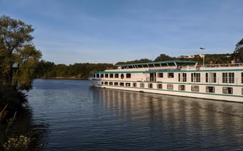Bateau de croisière MS Switzerland naviguant sur un fleuve calme avec des arbres verts sur la rive gauche et des bâtiments visibles au loin.