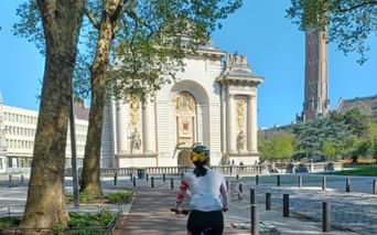 Cycliste se dirigeant vers la Porte de Paris, arc de triomphe blanc à Lille. Des arbres encadrent la scène sous un ciel bleu.