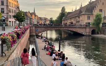Personnes se détendant au bord d'un canal à Strasbourg. Un pont en pierre traverse l'eau, bâtiments historiques et clochers en arrière-plan.